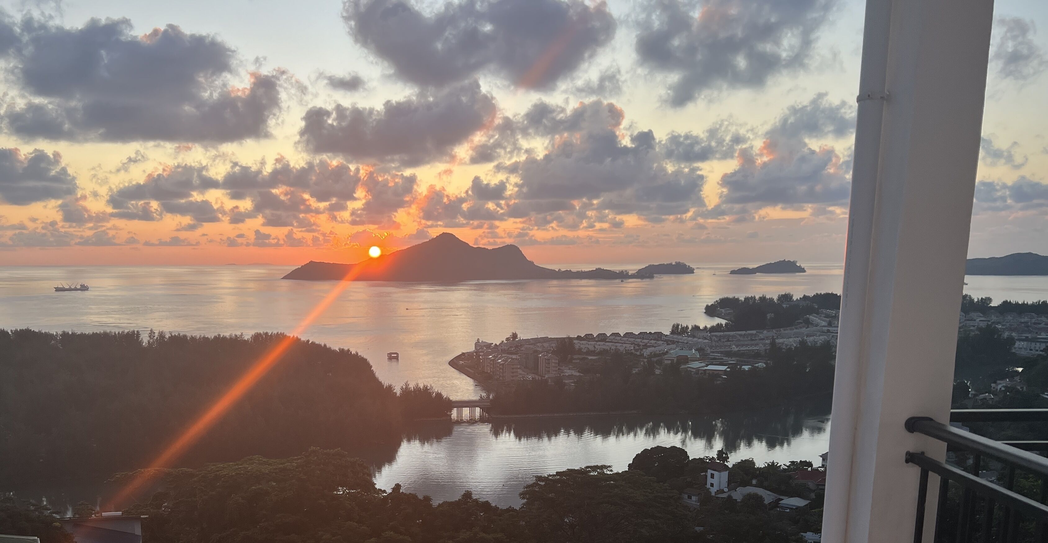 Vista do pôr do sol a partir de uma varanda com vista para águas calmas, ilhas distantes e uma ponte.