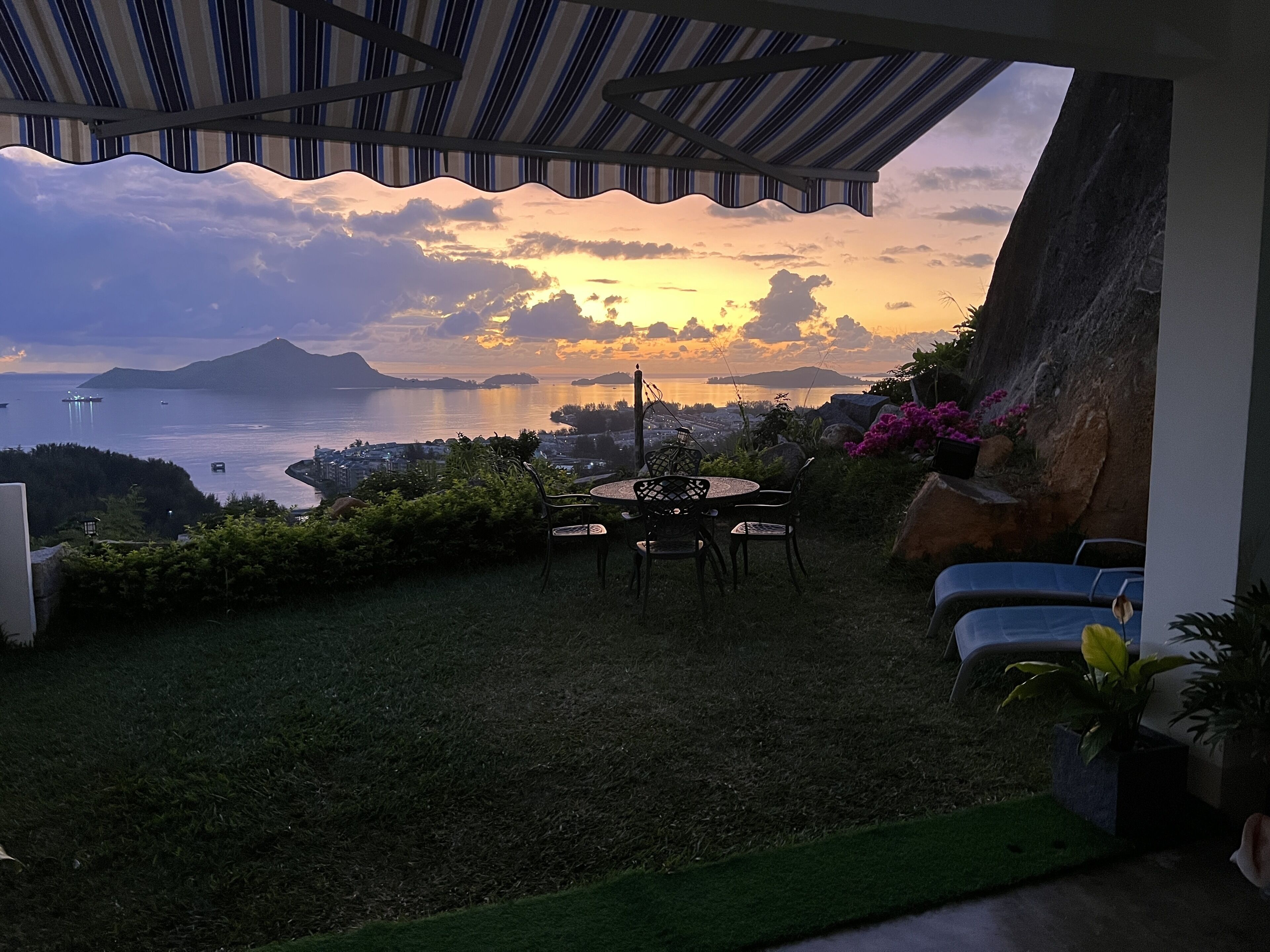 Un patio con mesa y sillas, con vistas panorámicas al agua y montañas distantes al atardecer.