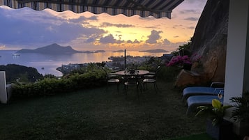 Un patio con mesa y sillas, con vistas panorámicas al agua y montañas distantes al atardecer.
