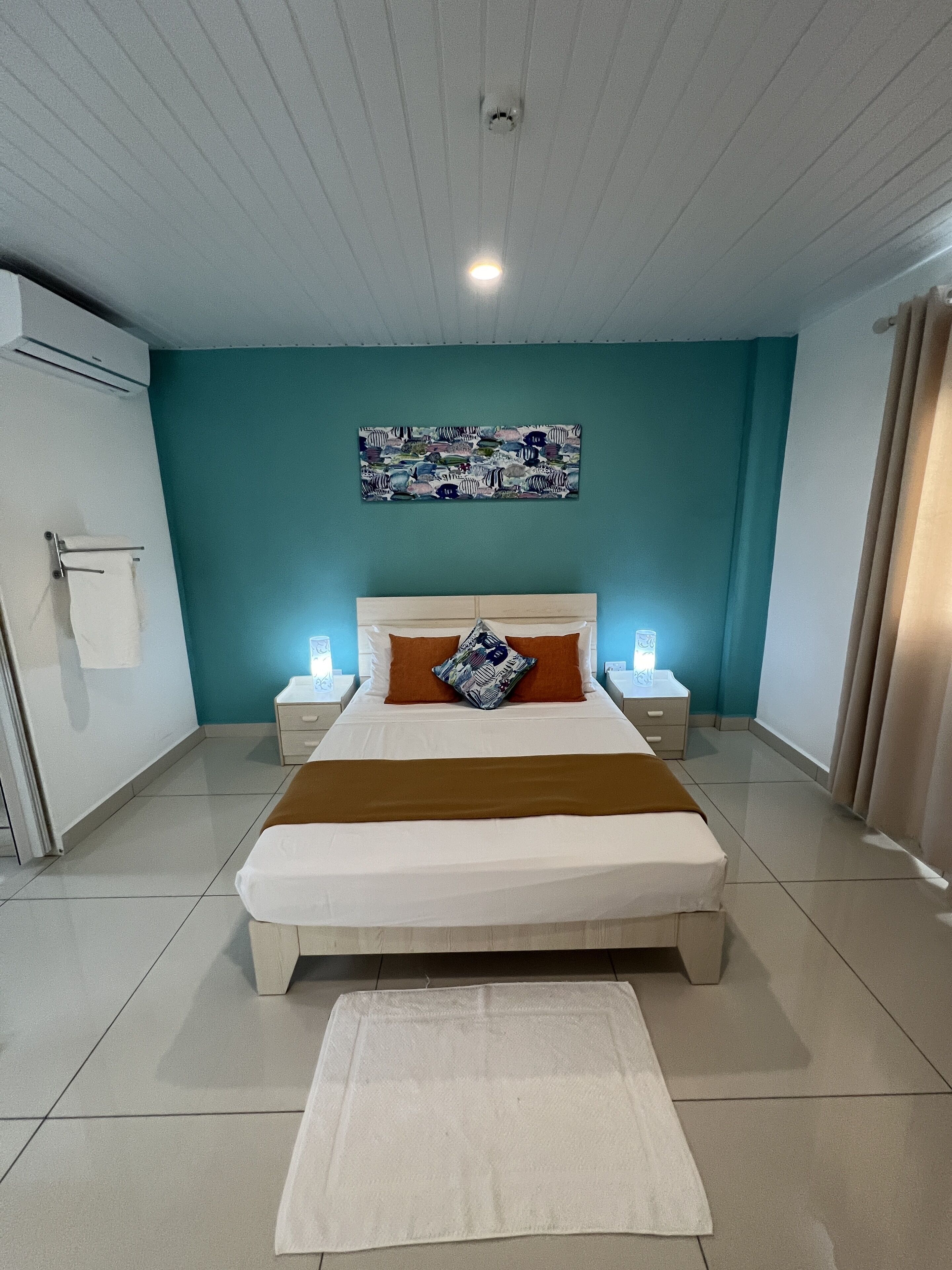 A modern bedroom with a large bed, a headboard, a nightstand, a wall-mounted air conditioner, a towel rack, and a ceiling light.