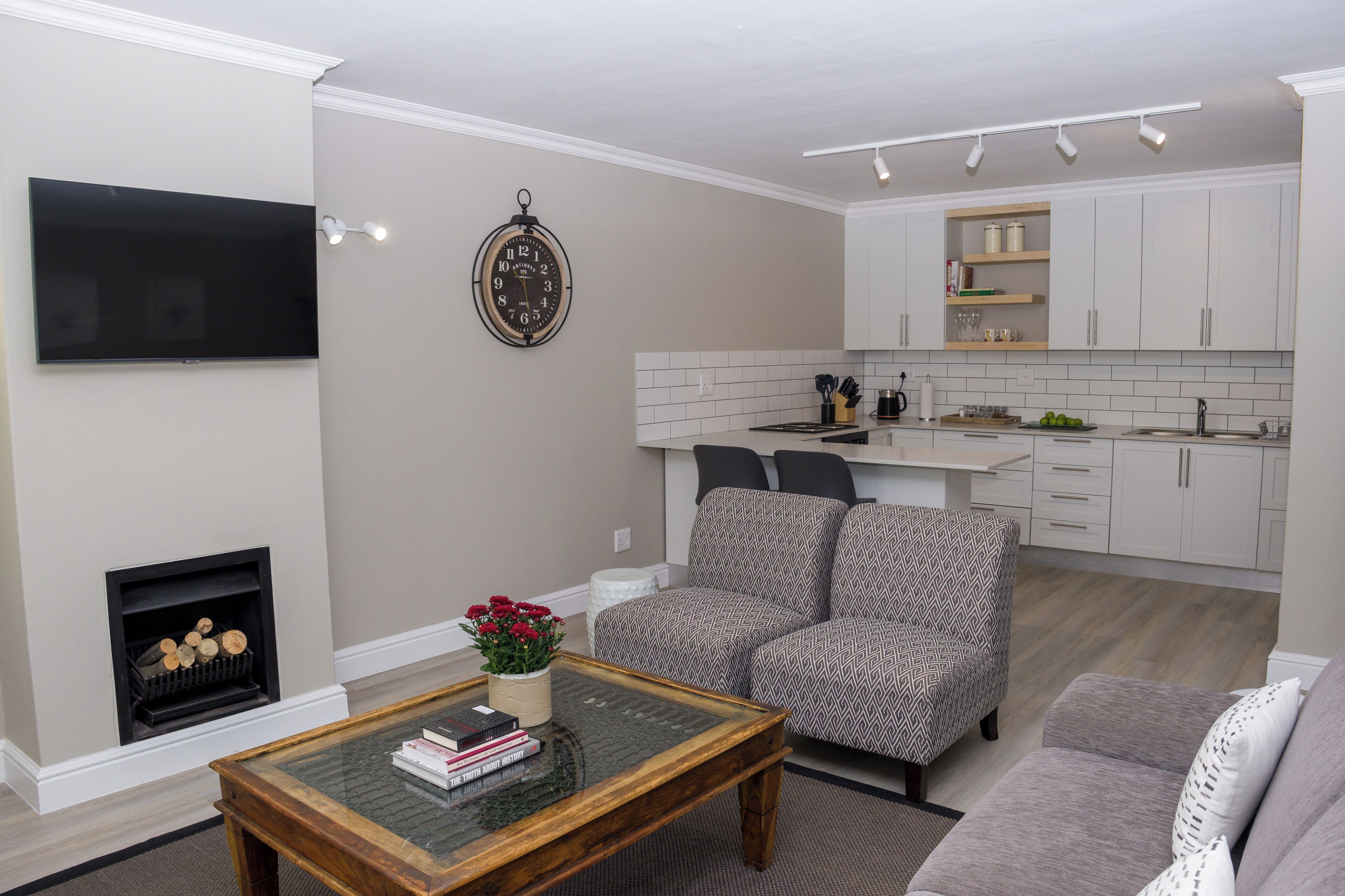 A living room with a fireplace, a television, a coffee table with books, patterned armchairs, and a kitchen area with cabinets and a countertop.