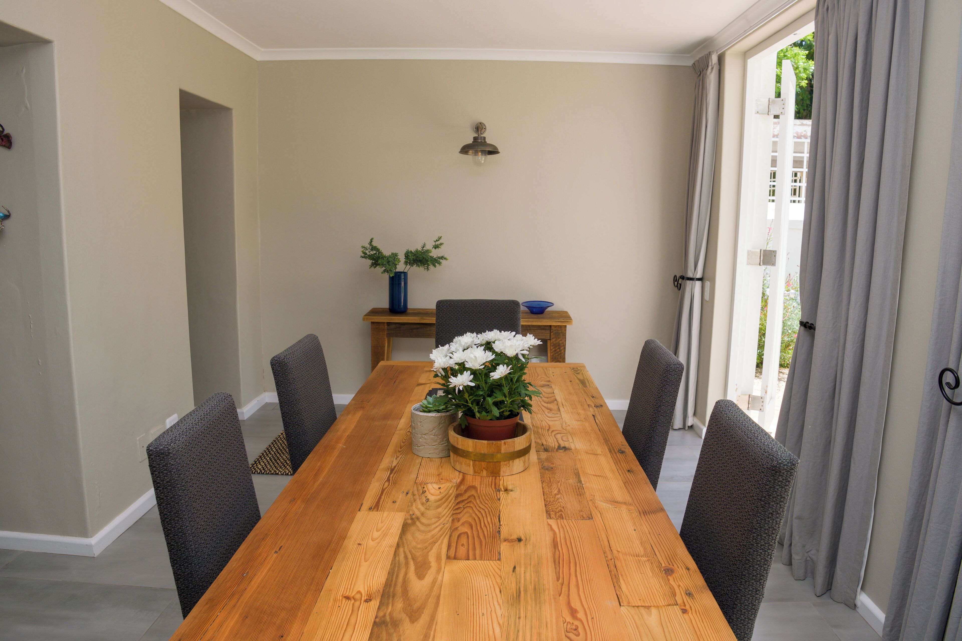 A dining room with a wooden table, six chairs, a vase with flowers, and a wall-mounted light fixture.