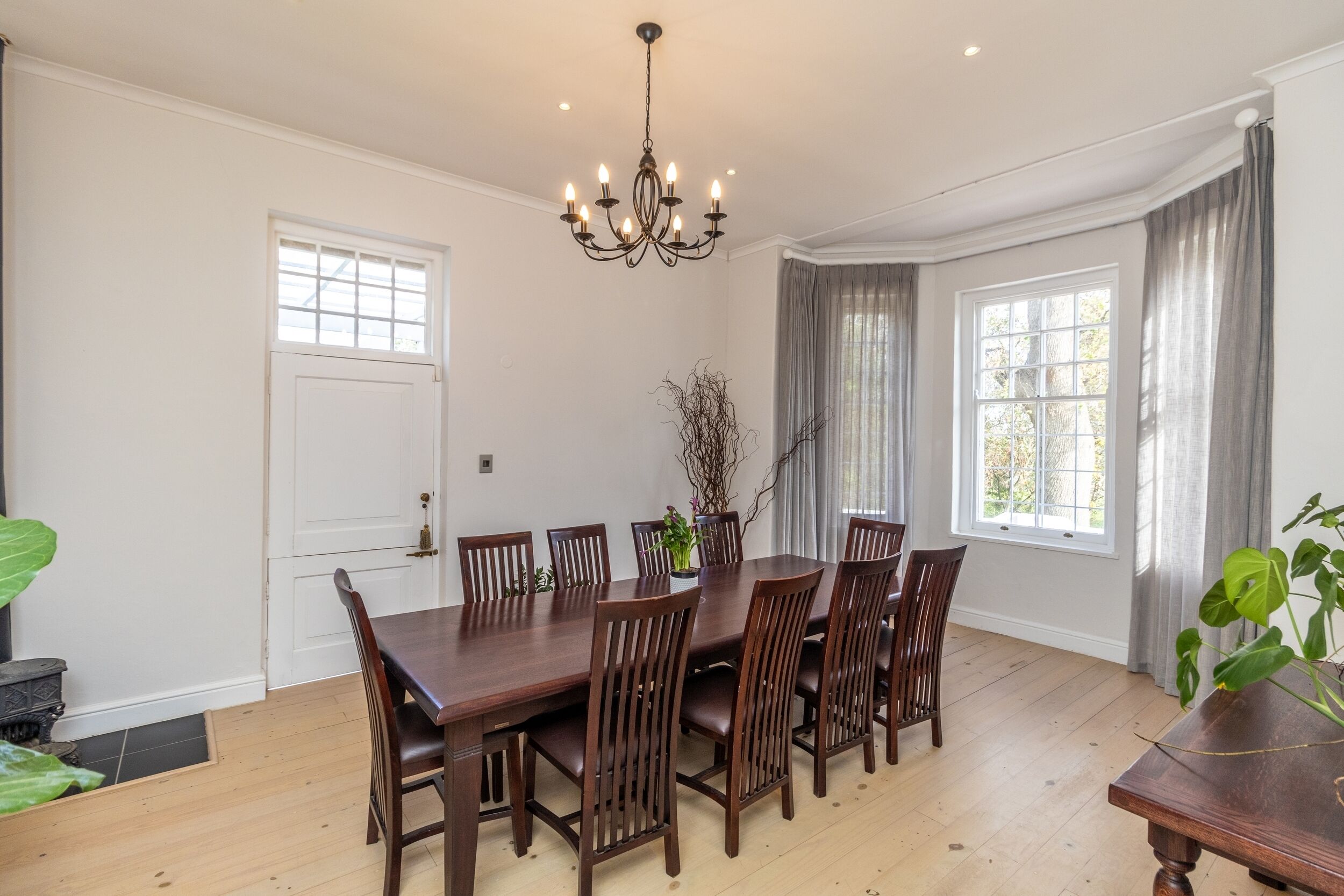 A dining room with a large wooden table and chairs, a chandelier, and windows with curtains.
