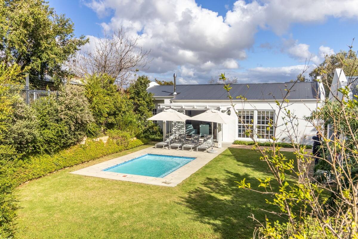 Un jardin avec une piscine, un espace salon couvert et une maison dotée d’une grande fenêtre donnant sur la piscine.