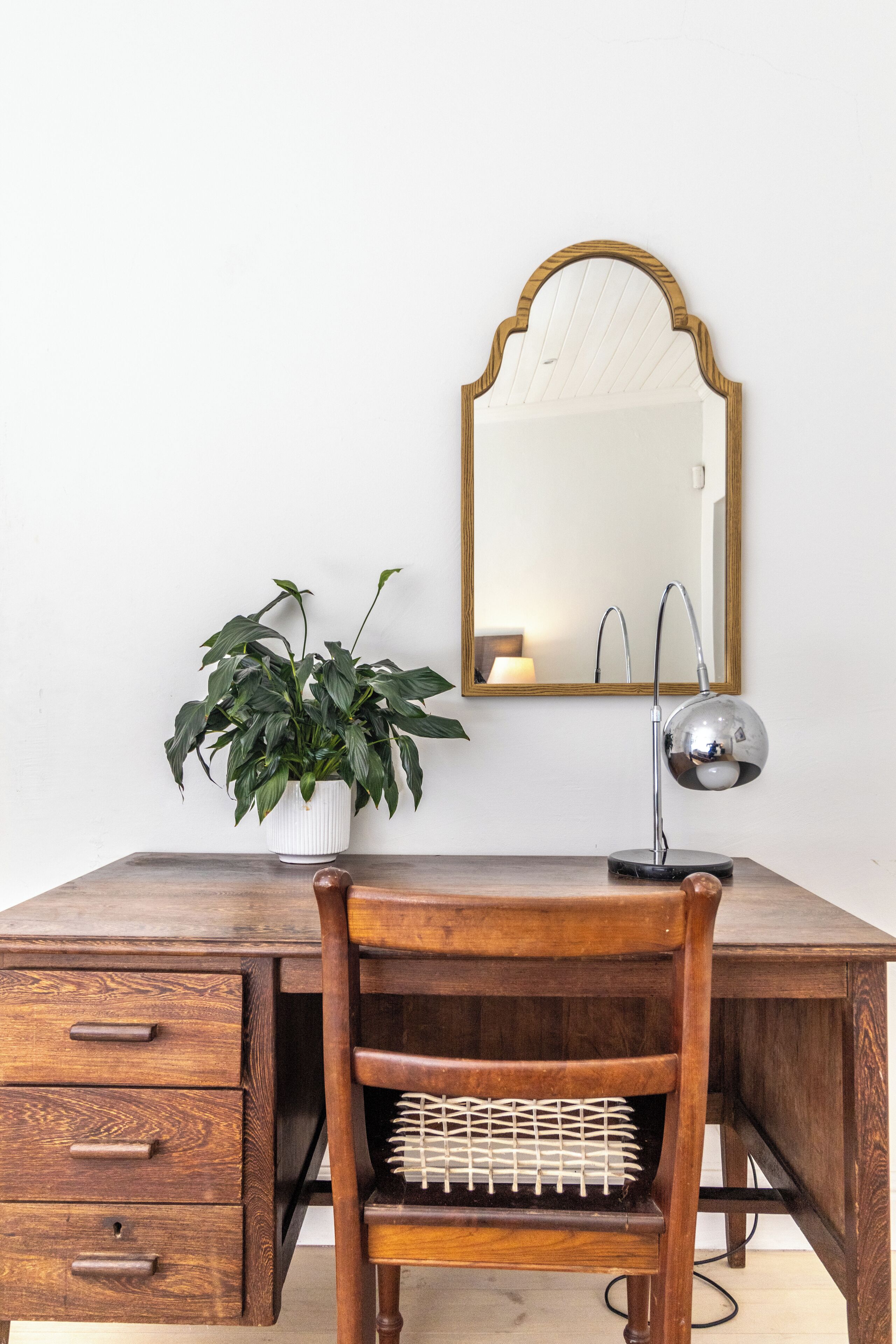 A wooden desk with a mirror, a potted plant, and a chair with a woven seat.