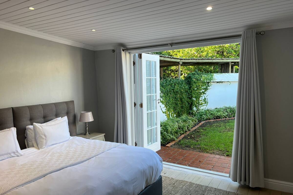 Une chambre avec un grand lit, une table de chevet avec une lampe, et une vue sur une piscine et un jardin, grâce à une porte ouverte.