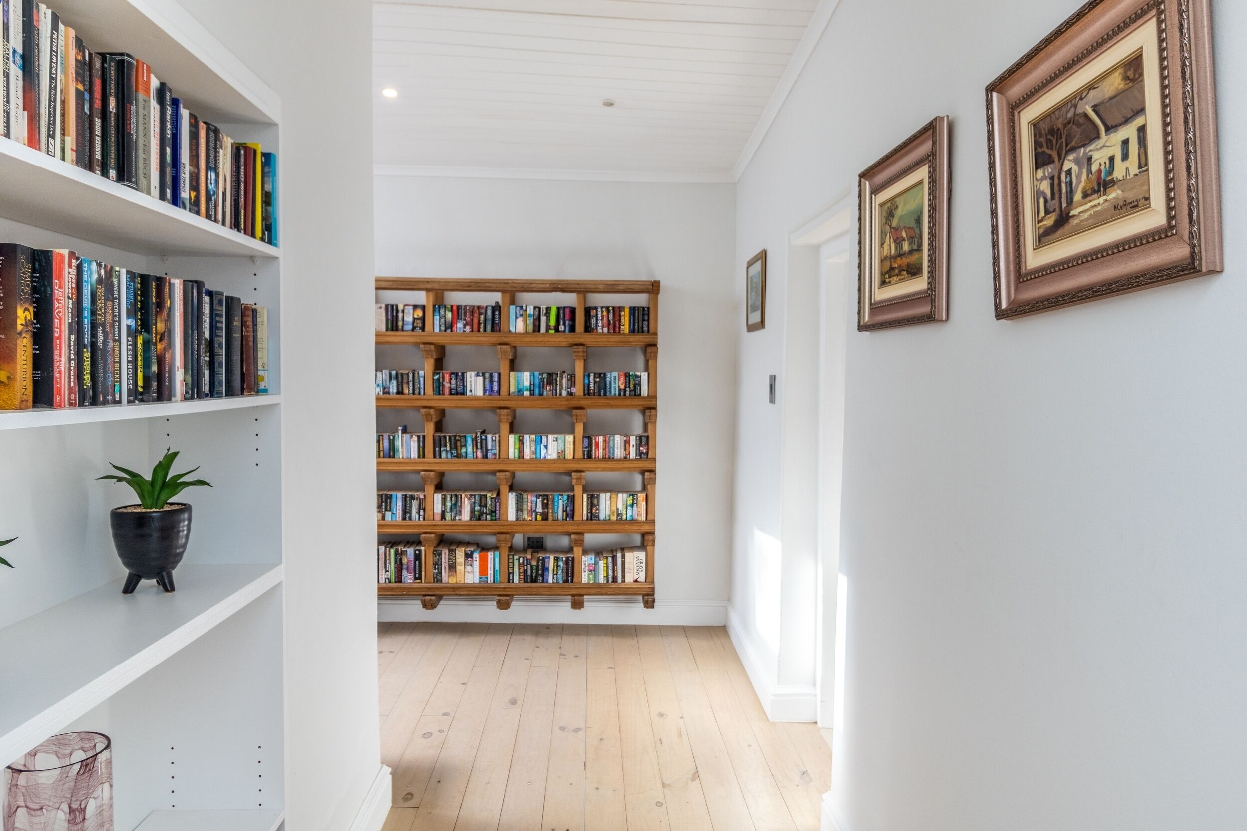 A hallway with white shelves on the left, a large wooden bookshelf in the center, and framed pictures on the right wall.