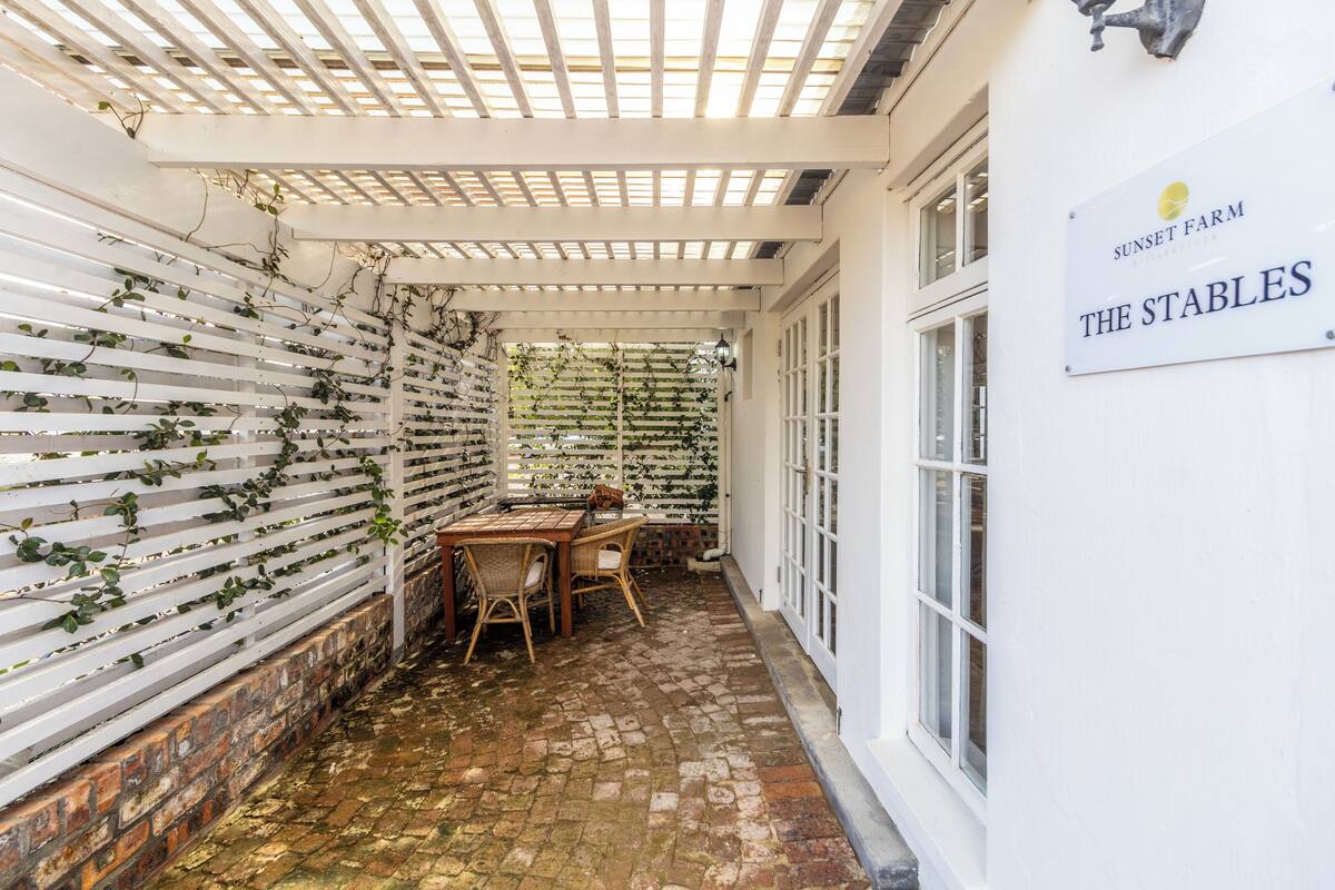 Un espace extérieur abrité, doté d’un sol en briques, d’un mur en treillis blanc et d’une table avec des chaises. Une enseigne sur le mur indique : « Sunset Farm, les écuries ».