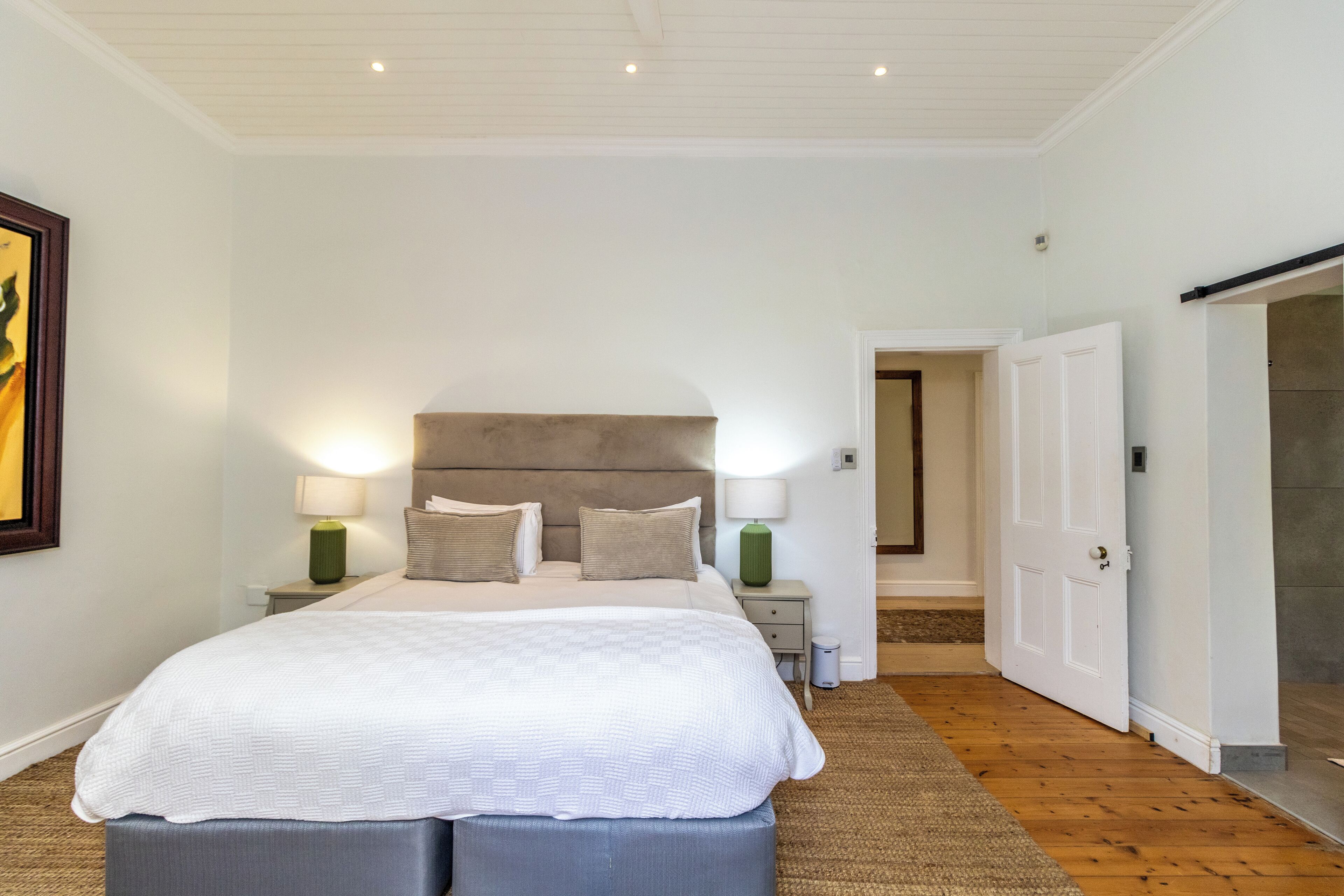 Une chambre avec un grand lit, deux tables de chevet avec des lampes, une porte donnant sur une autre pièce et un tableau encadré au mur.