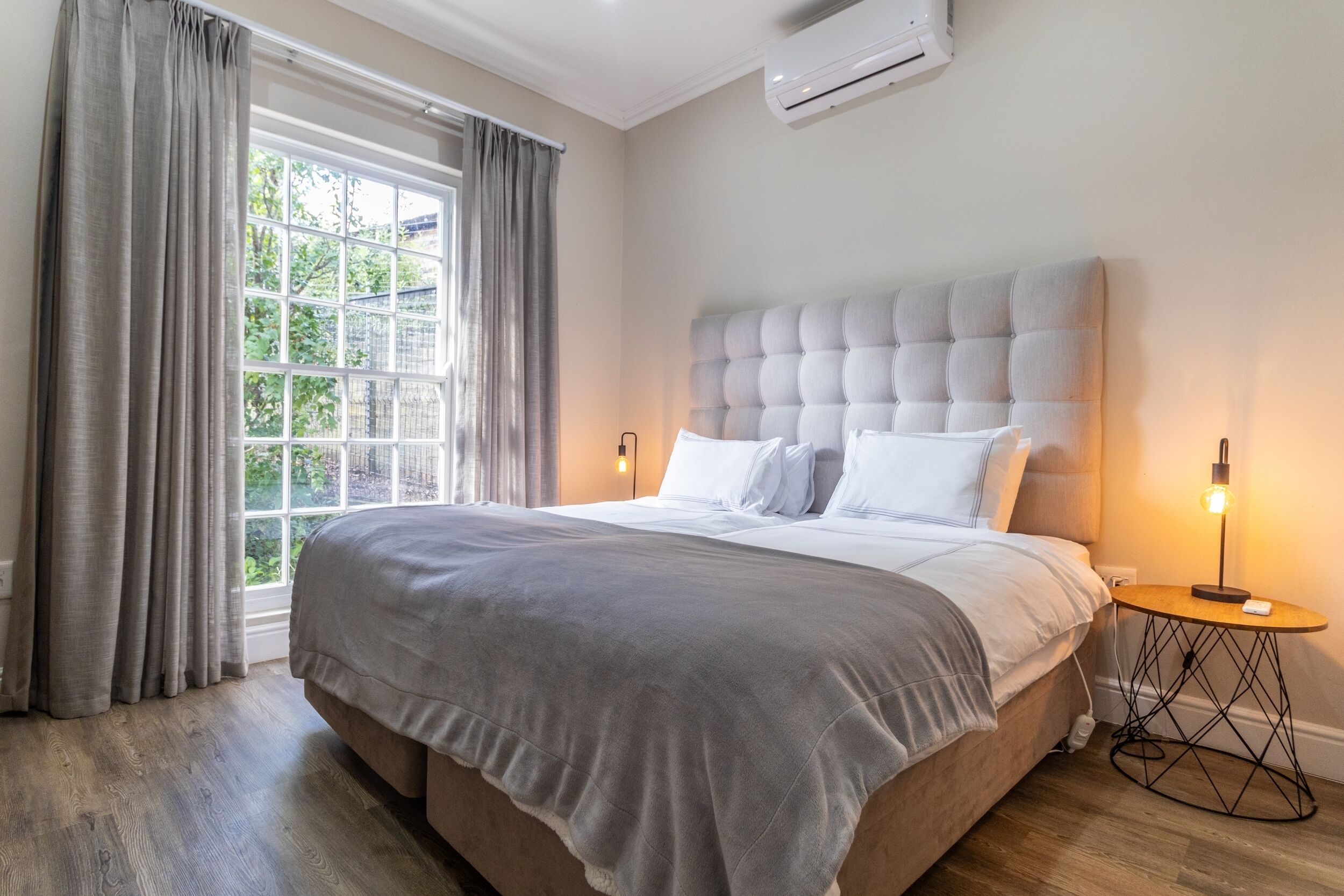 A bedroom with a large bed, a window with curtains, a bedside lamp, and an air conditioning unit.