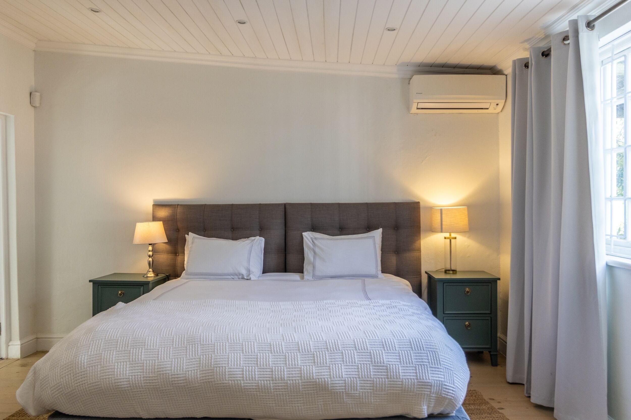 Een slaapkamer met een groot bed, twee nachtkastjes met lampen, een wandairconditioner en een raam met gordijnen.