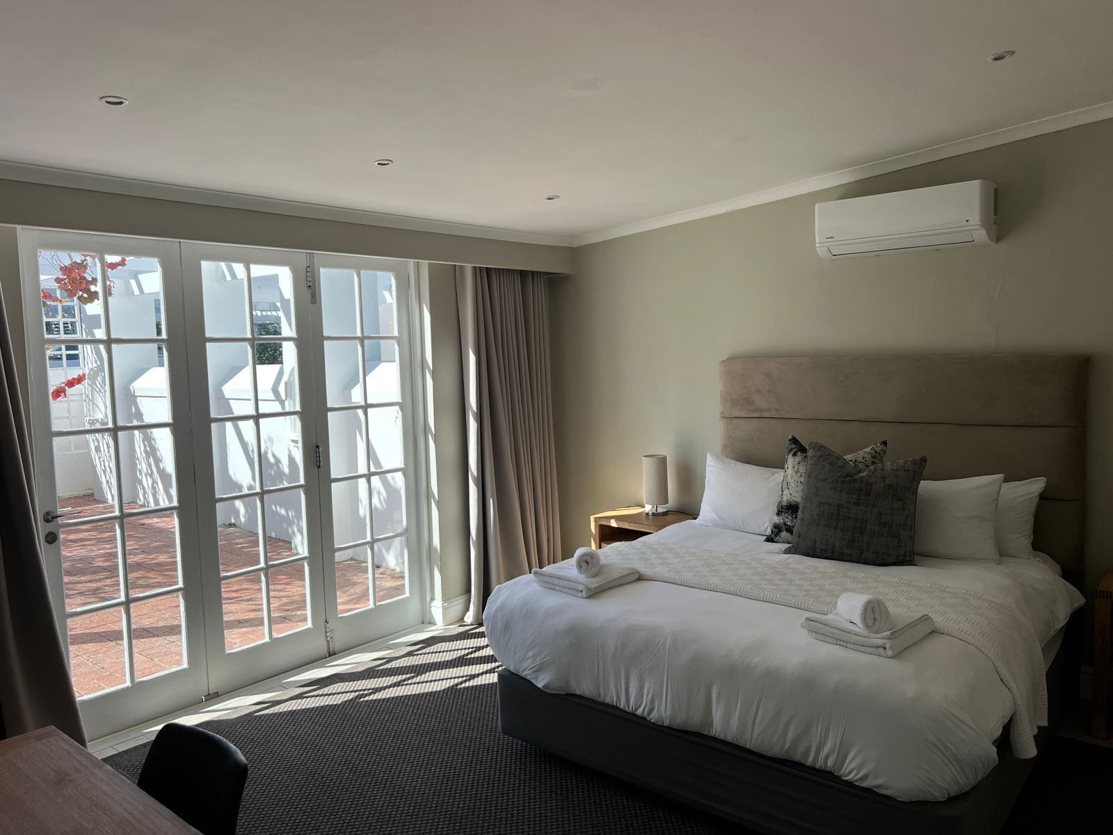 A hotel room with a large bed, a headboard, a nightstand, an air conditioning unit, and a view of a courtyard with outdoor furniture.