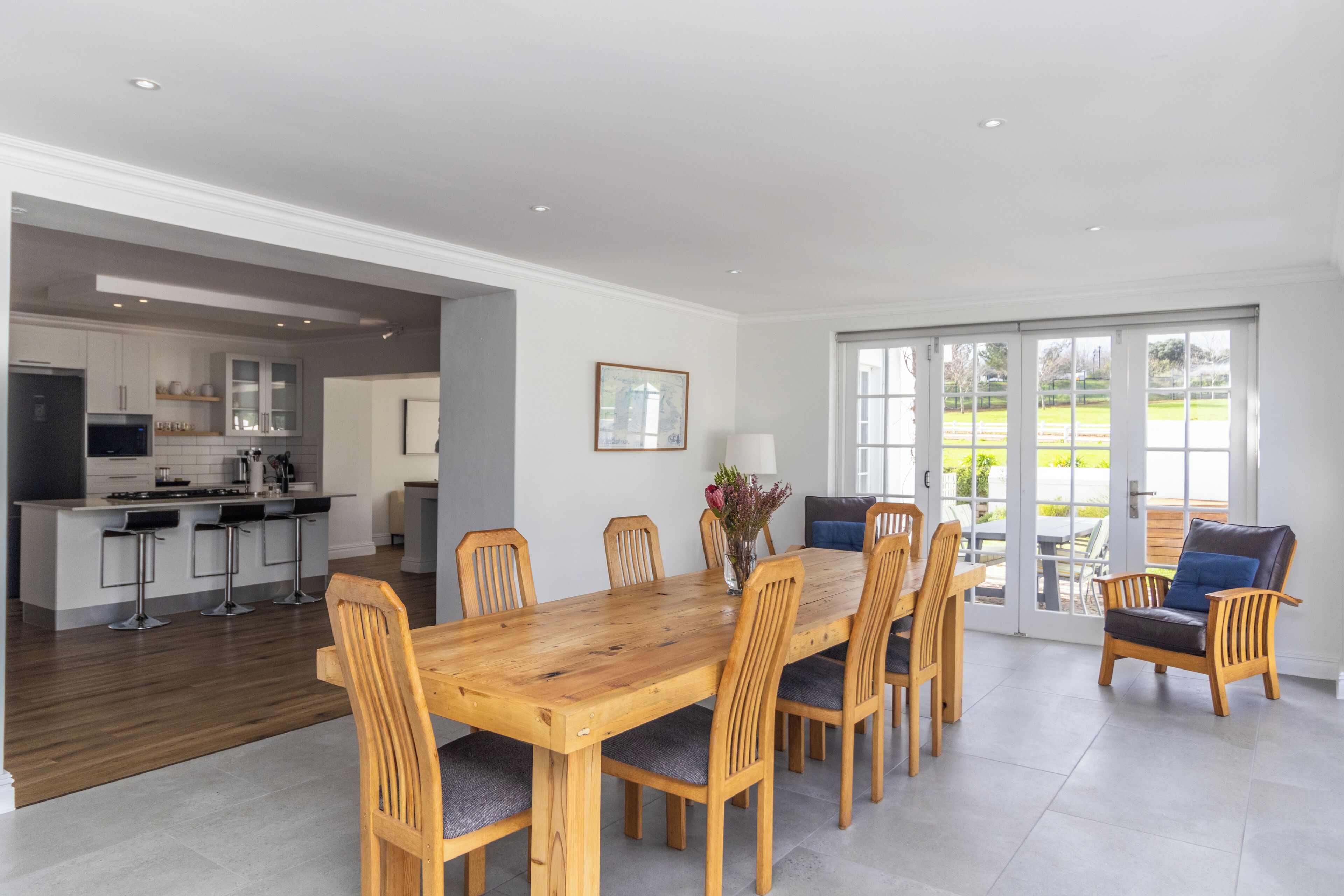 A spacious dining area with a wooden table and chairs, a kitchen in the background, and a view through large glass doors.