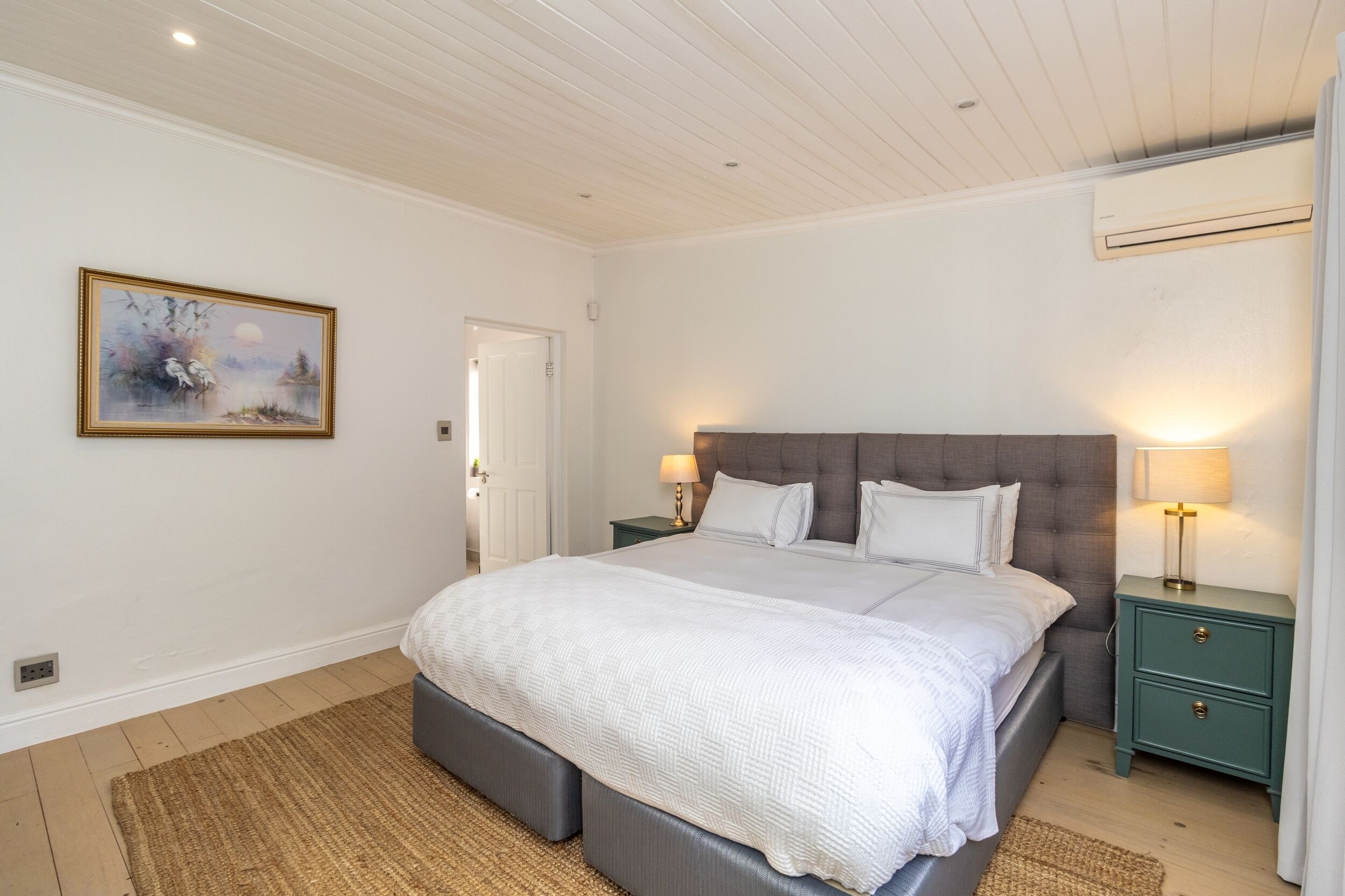 A bedroom with a large bed, two bedside tables with lamps, a painting on the wall, and a door leading to another room.