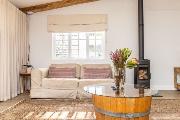 A cozy living room with a beige sofa, a glass coffee table, a fireplace, and a window with curtains. - Sunset Farm Stellenbosch (Stellenbosch)