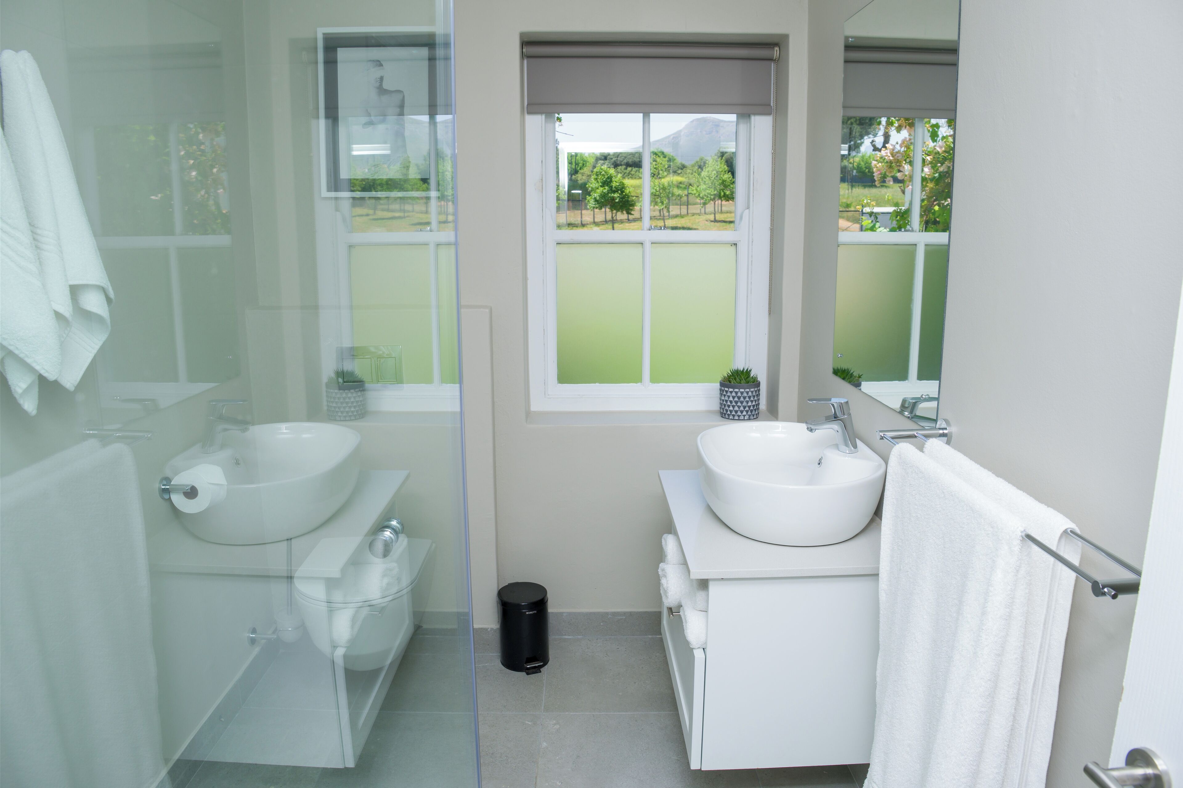 Un baño moderno con cabina de ducha de vidrio, dos lavamanos y un amplio ventanal con vistas a zonas verdes y un edificio.