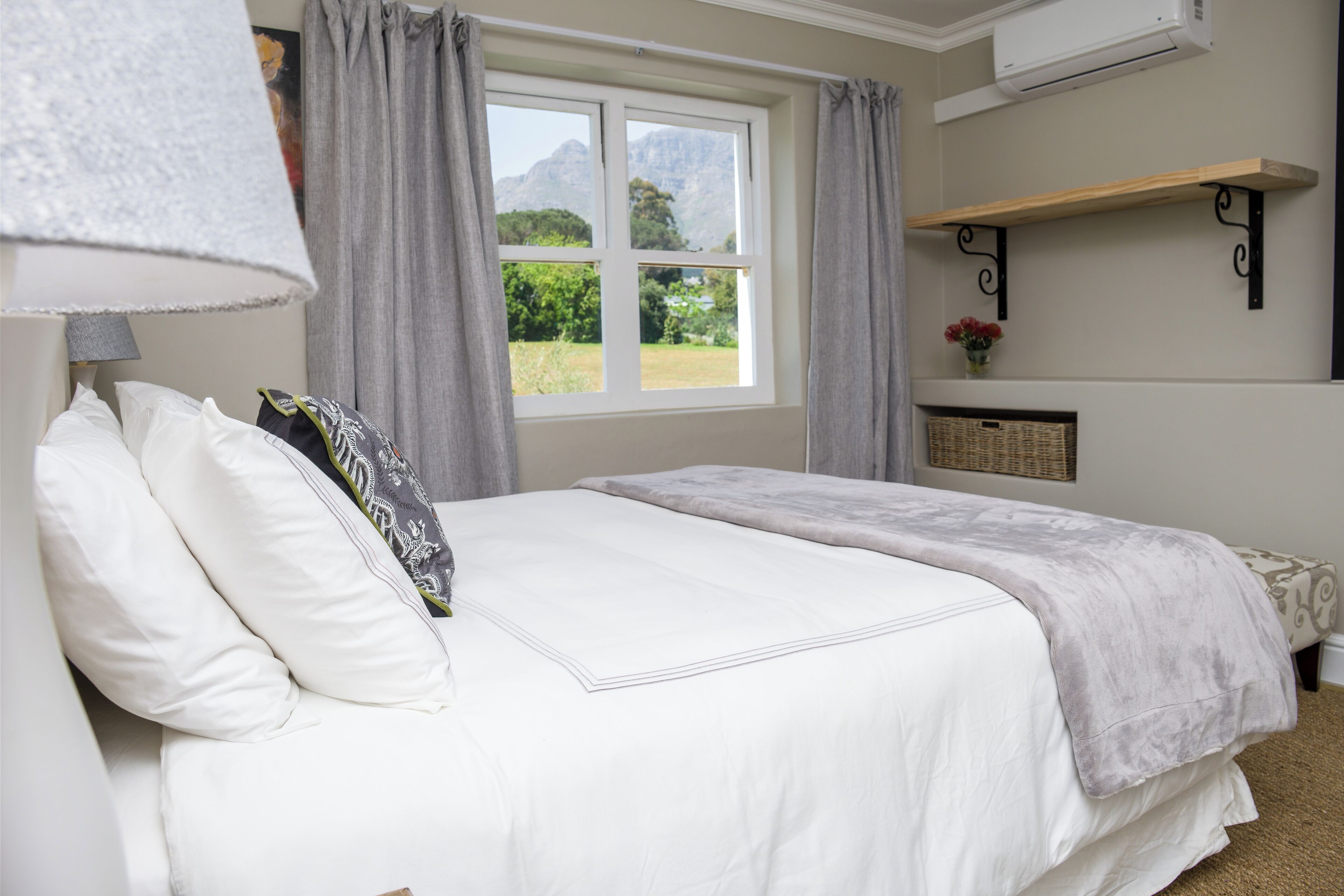 A bedroom with a bed, a window with curtains, a wall-mounted shelf with a basket, and an air conditioning unit.