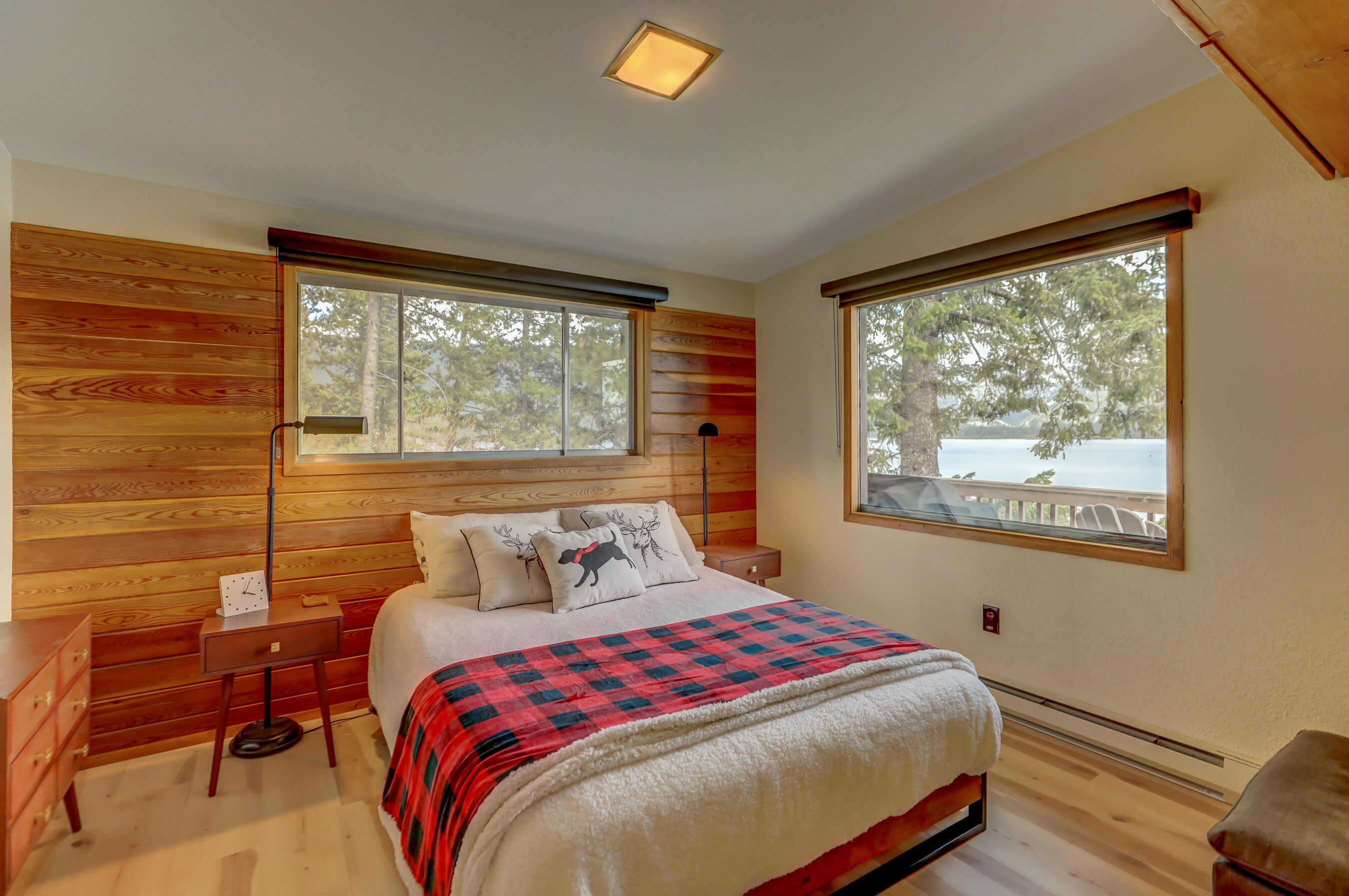 Une chambre à coucher avec une tête de lit en bois, un lit recouvert d’une couverture à carreaux rouges et noirs, deux tables de chevet, une commode et de grandes fenêtres donnant sur des arbres et un balcon.