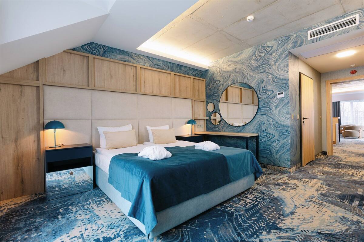 Une chambre d’hôtel avec un grand lit, des boiseries, un miroir rond et une moquette à motifs.