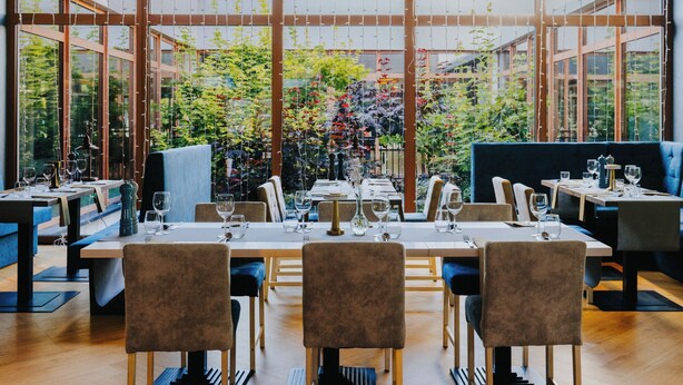A restaurant with a long table set for dining, surrounded by chairs, and large windows offering a view of greenery outside. - Aspen Prime Ski & Bike Resort (Podlesie)
