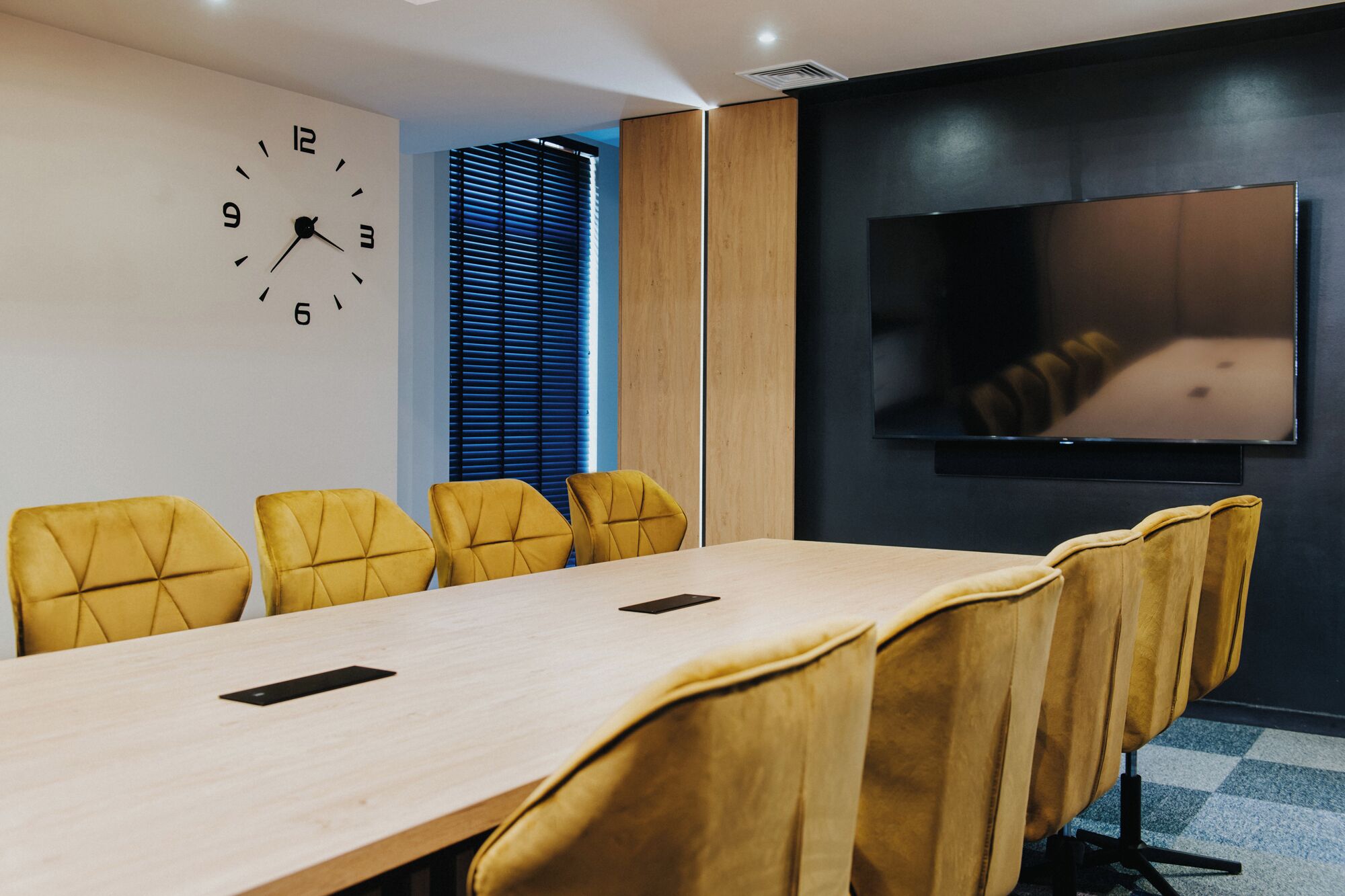 A conference room with a long table, yellow chairs, a clock, and a TV on the wall.