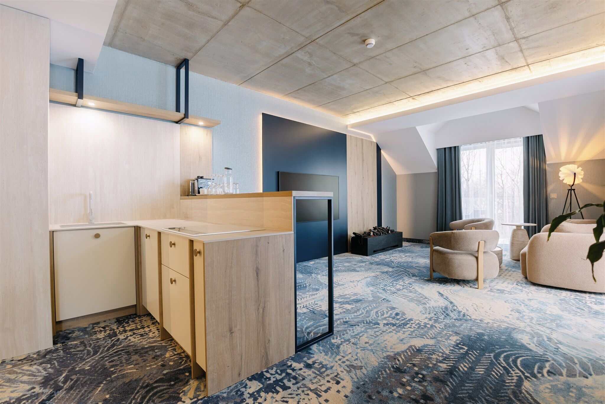 Une chambre d’hôtel moderne dotée d’une kitchenette, d’une grande fenêtre avec des rideaux, d’un tapis à motifs et d’un coin salon avec un canapé et des fauteuils.