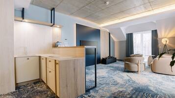 Une chambre d’hôtel moderne dotée d’une kitchenette, d’une grande fenêtre avec des rideaux, d’un tapis à motifs et d’un coin salon avec un canapé et des fauteuils.