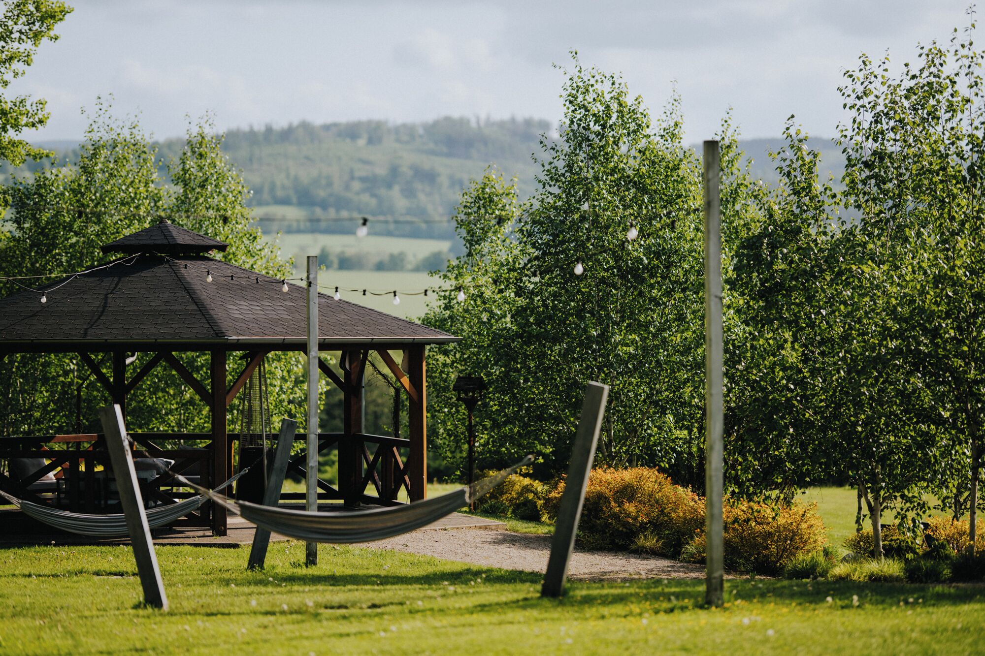 A gazebo with a hammock, surrounded by greenery and a view of hills.