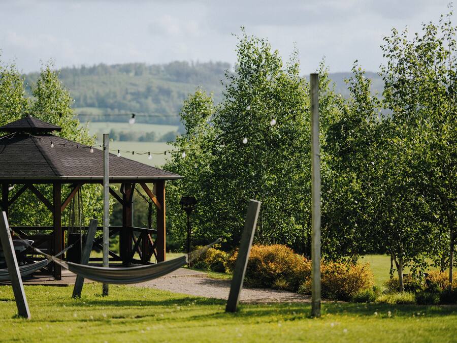 A gazebo with a hammock, surrounded by greenery and a view of hills.