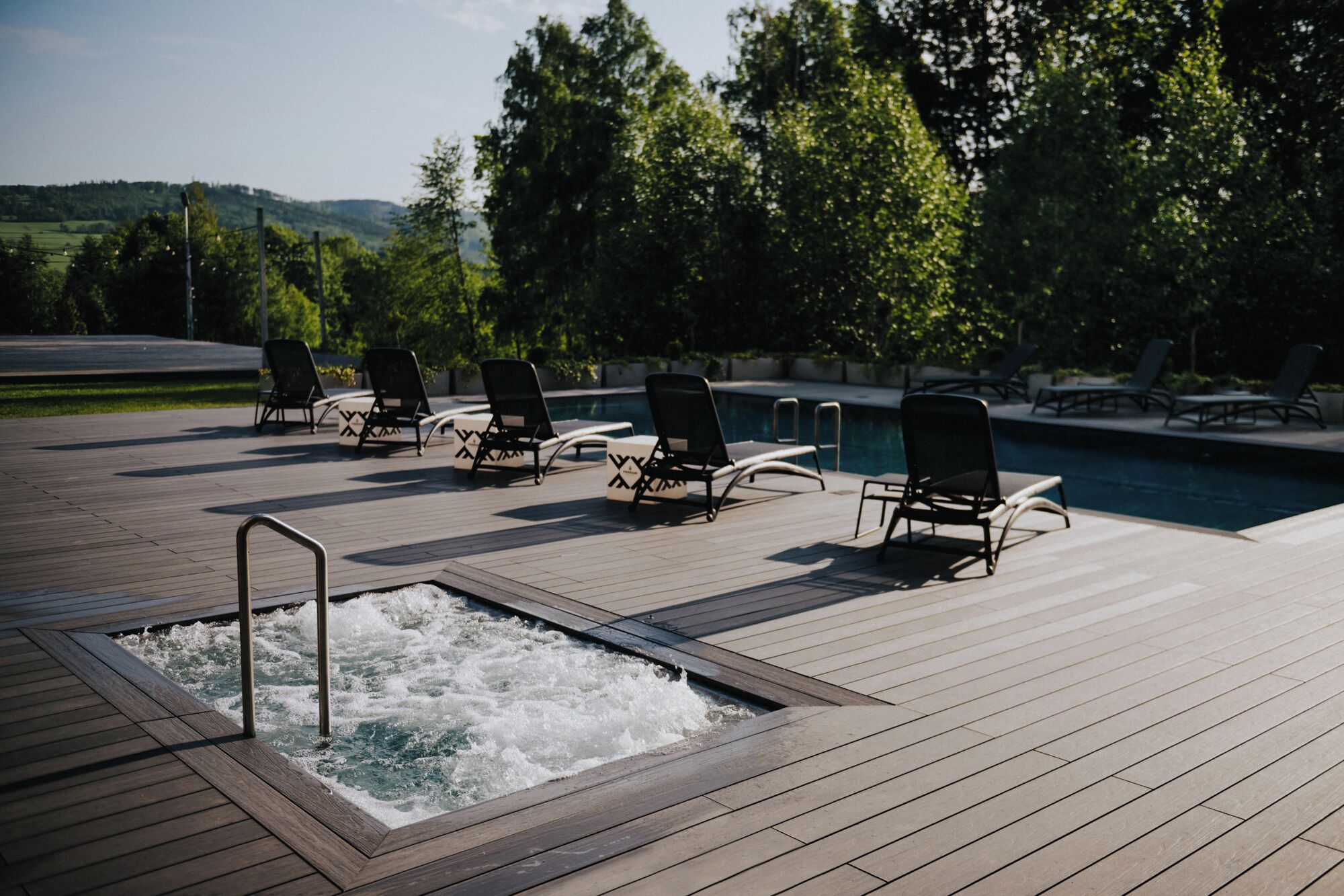 A wooden deck with a hot tub and lounge chairs, overlooking a swimming pool and a scenic landscape.