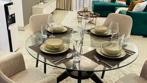 Un salon moderne avec une table à manger en verre, préparée pour quatre personnes, entourée de fauteuils beiges. Un canapé vert et un meuble blanc avec un téléviseur à écran plat se trouvent à l’arrière-plan.