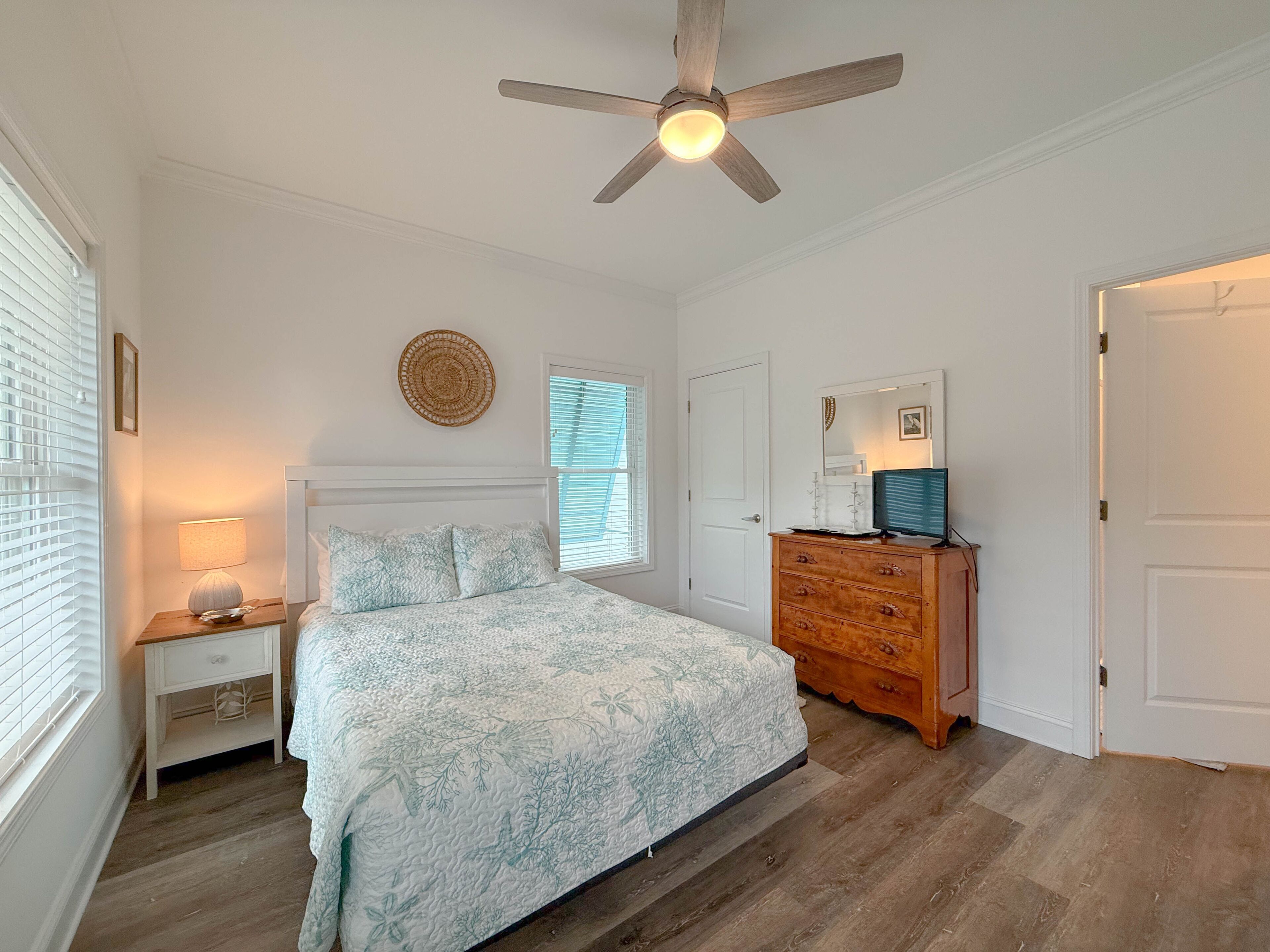 A bedroom with a bed, a dresser, a ceiling fan, a window with blinds, and a wall-mounted round mirror.