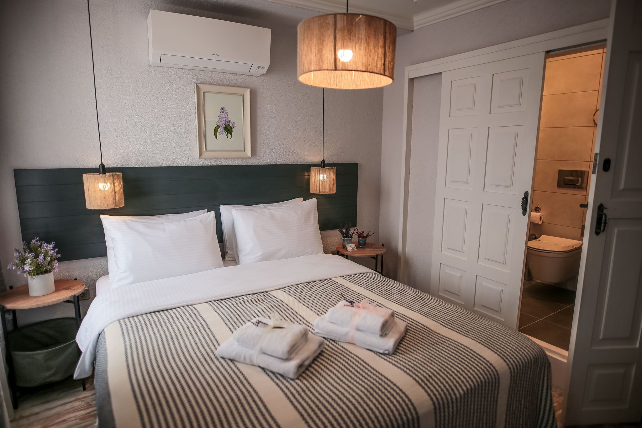 Une chambre d’hôtel avec un grand lit, deux tables de chevet, un climatiseur fixé au mur, un tableau encadré et une porte donnant accès à la salle de bain.