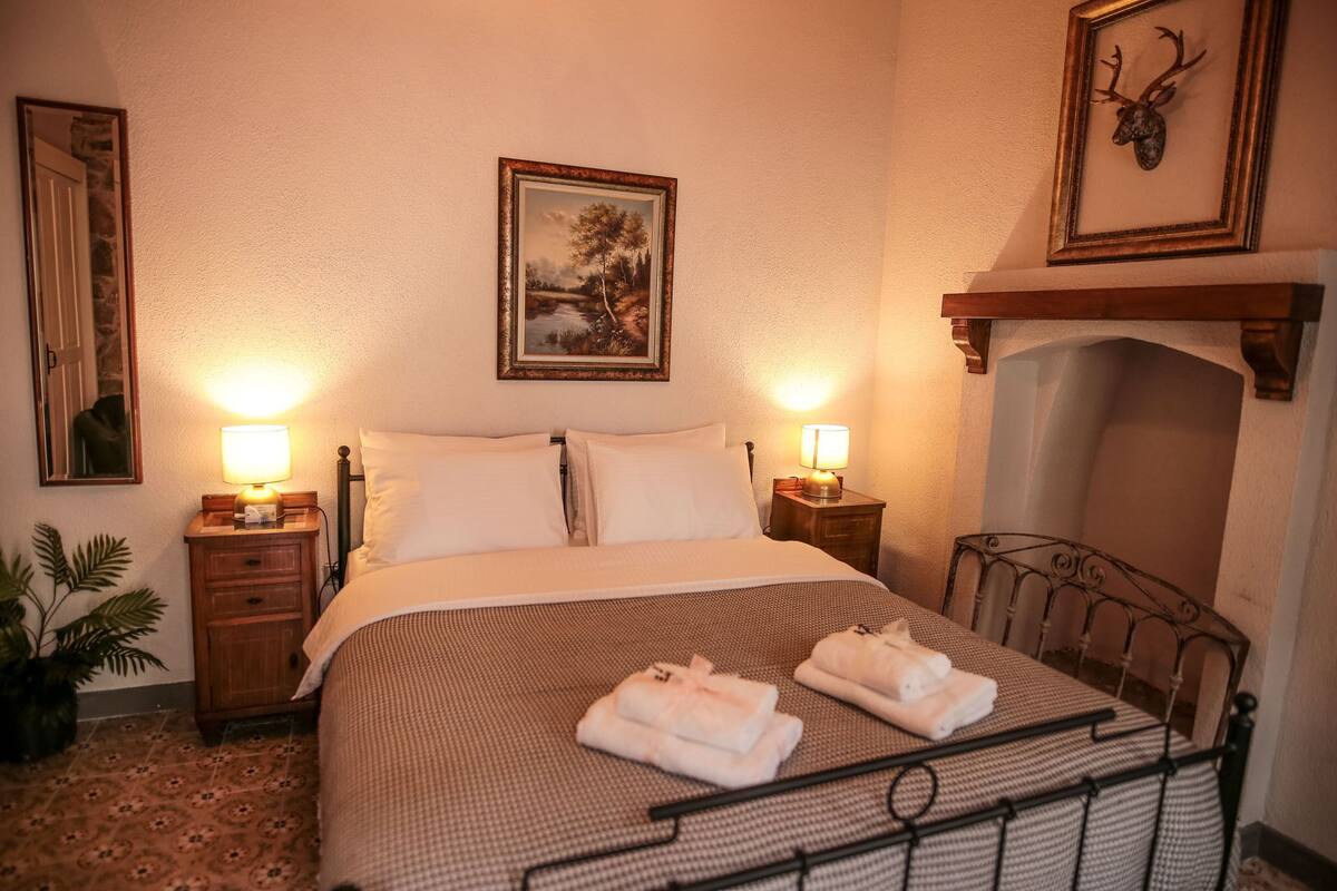 A neatly made bed with a checkered bedspread, two bedside lamps, a framed picture on the wall, and a mounted deer head above a fireplace.