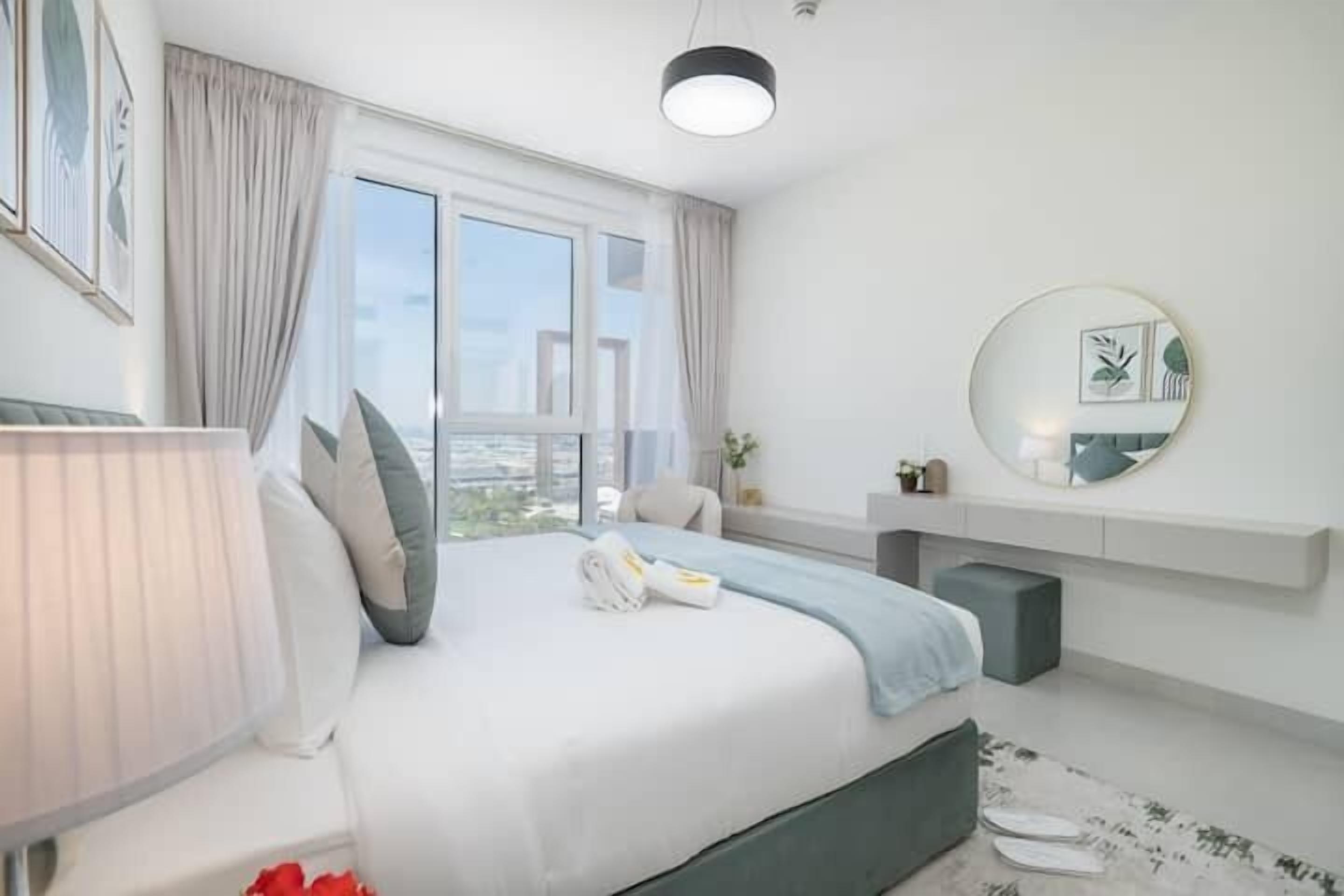 A modern hotel room with a large bed, a bedside table, a lamp, a round mirror, and a view of the city through the window.