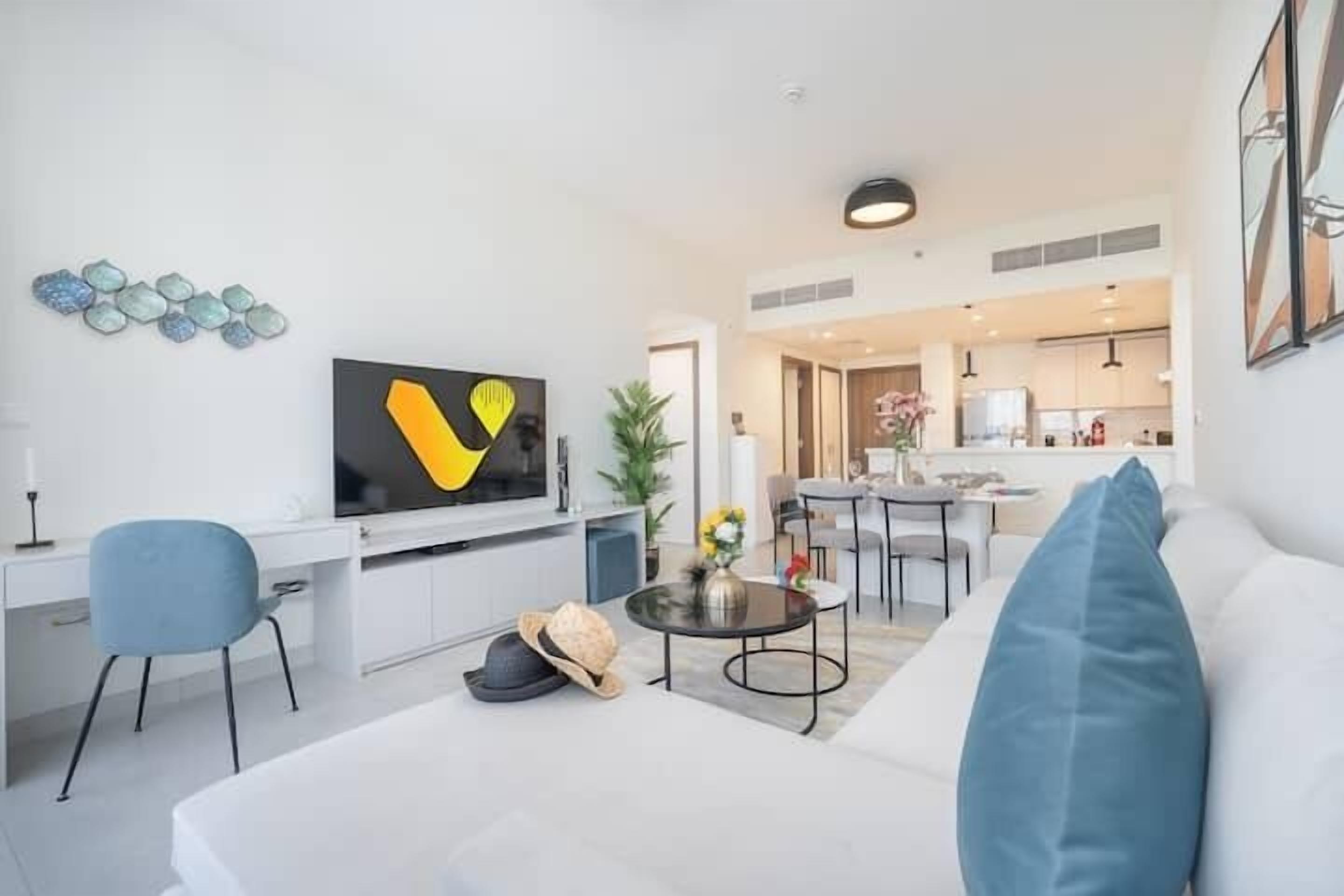 A modern living room with a sofa, a TV, a dining area, and a kitchen in the background.