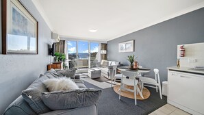 Ein modernes Wohnzimmer mit einem grauen Sofa, einem Esstisch mit Stühlen und einer Küchenzeile mit einem weißen Geschirrspüler.