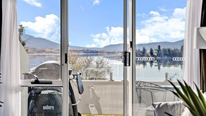 Blick durch eine Glastür auf einen Raum mit einem großen Fenster mit Blick auf einen See, Berge und eine Brücke.