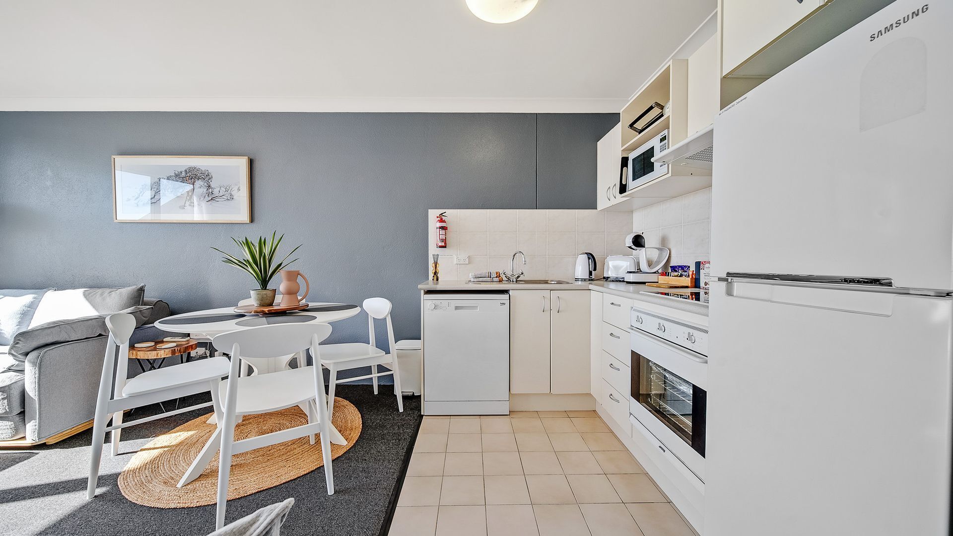 A modern kitchen with white cabinetry, a dishwasher, and a microwave. There is a dining area with a table and chairs, and a small rug on the floor.