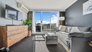 Ein modernes Wohnzimmer mit einer Couch, einem Holzschrank, einem Fernseher und einem großen Fenster mit Blick ins Freie.