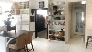 Ett modernt kök med vita skåp, en matplats med flätade stolar och en inbyggd hyllsektion med olika köksredskap.