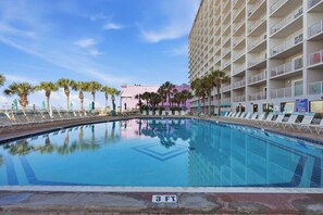 A pool with lounge chairs, palm trees, and a hotel building in the background.