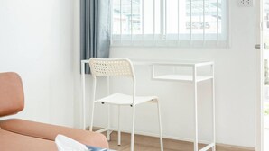 A room with a window, a white chair, a table with a tissue box and remote, and a brown couch. - Charming Studio Suite - Private Washer  (Chalong)