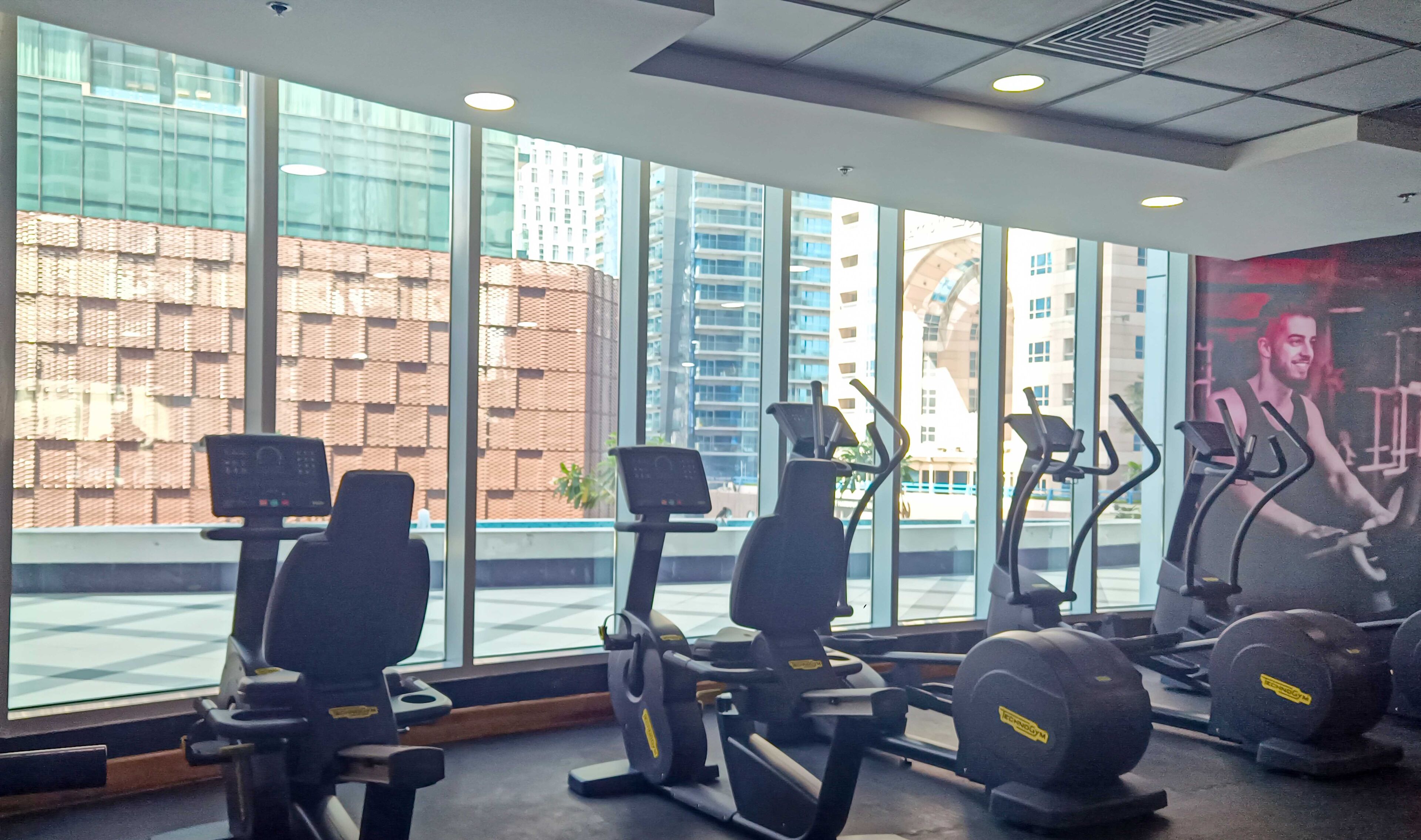A gym with exercise equipment and large windows overlooking a brick building.