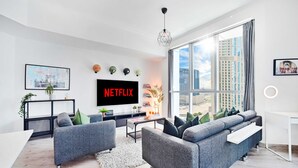 A modern living room with grey sofas, a large TV displaying 'NETFLIX', a city view through large windows, and a minimalist decor.