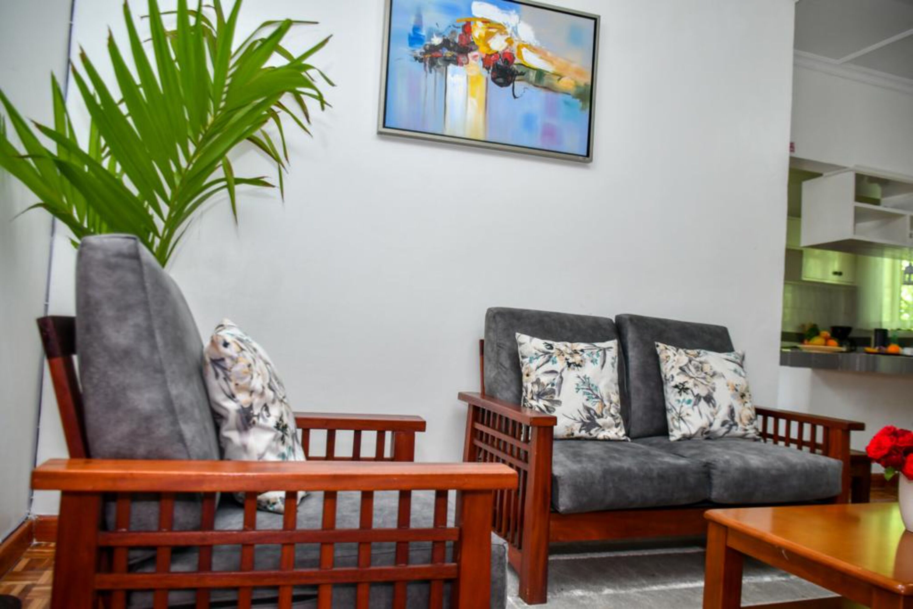 Ein Wohnzimmer mit grauem Sofa, Holzsesseln, einer großen Grünpflanze und einem farbenfrohen abstrakten Gemälde an der Wand.