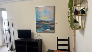 Un salon avec un téléviseur posé sur un meuble noir, un tableau accroché au mur et une plante sur une étagère.