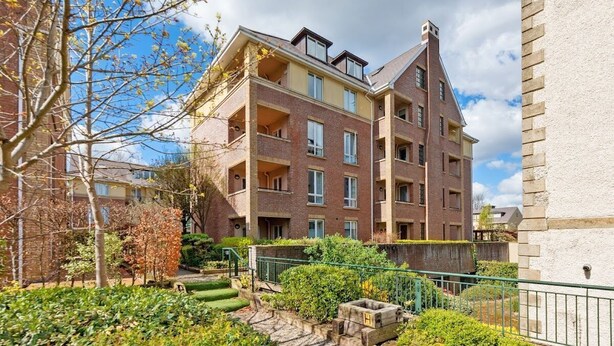 Un immeuble résidentiel à plusieurs étages, doté d’une façade en briques, de nombreux balcons et d’un petit jardin avec des marches menant à l’entrée.