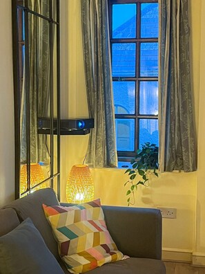 A living room with a sofa, a patterned throw pillow, a floor lamp, and a window with curtains.