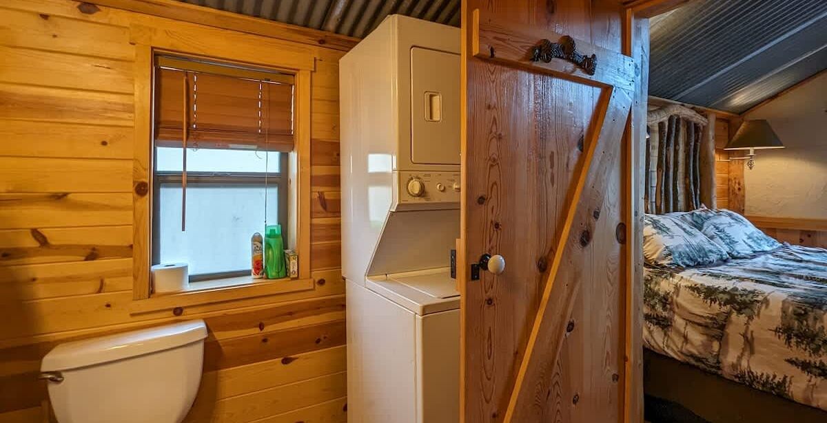 A wooden cabin interior with a toilet, washing machine, and a bed with a forest-themed bedspread.